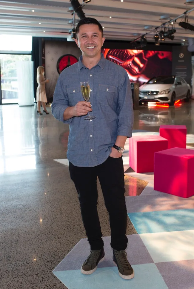 Man in casual wear holds a champagne glass at an indoor event, with a car display in the background.