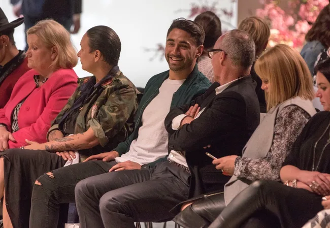 Group of people sitting in a row, some engaged in conversation, at an indoor event with floral decorations in the background.