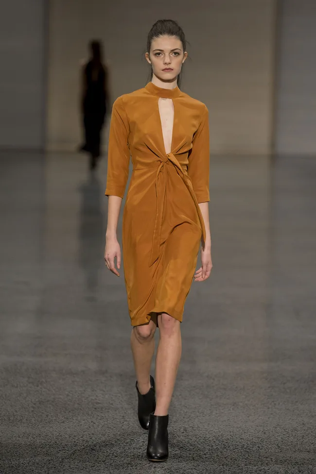 Model walking on a runway wearing a mustard yellow dress with cut-out detail and black ankle boots.