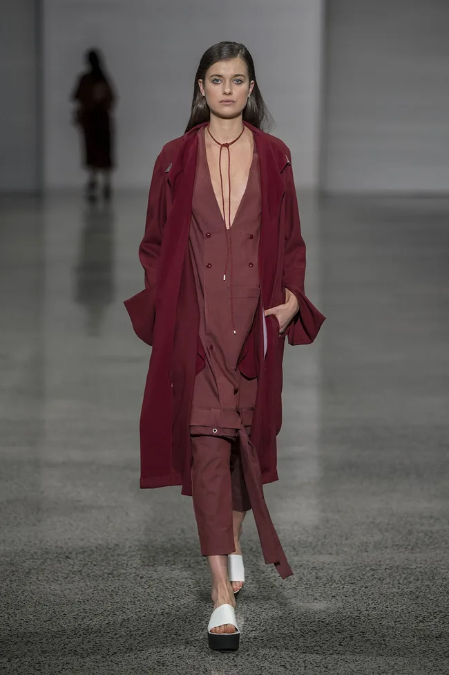 Model in a burgundy outfit and white sandals walking on a runway with gray background.