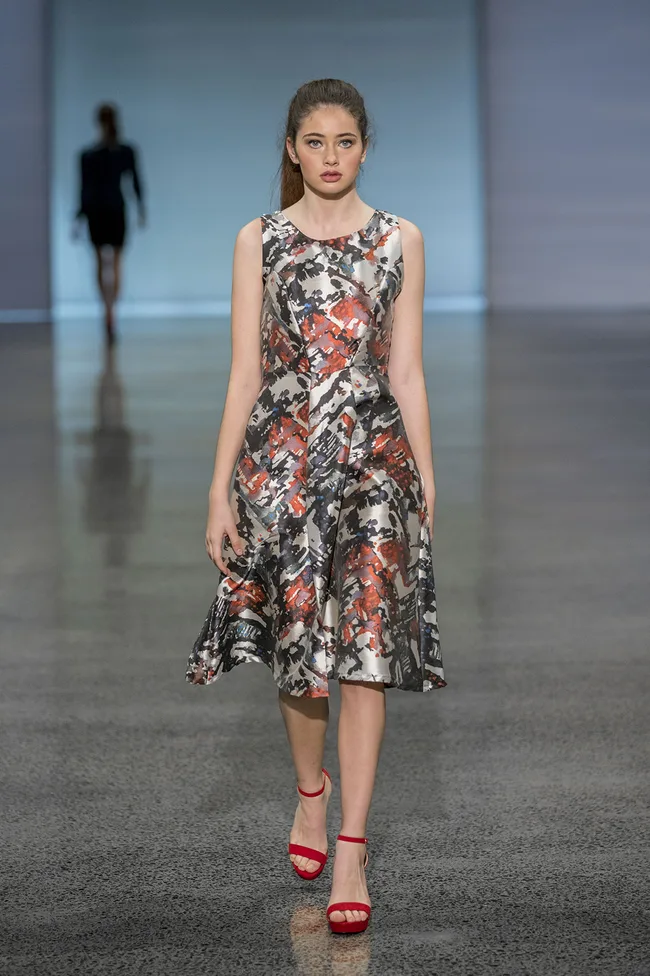 Model in a patterned dress and red heels walks down the runway, with another model blurred in the background.