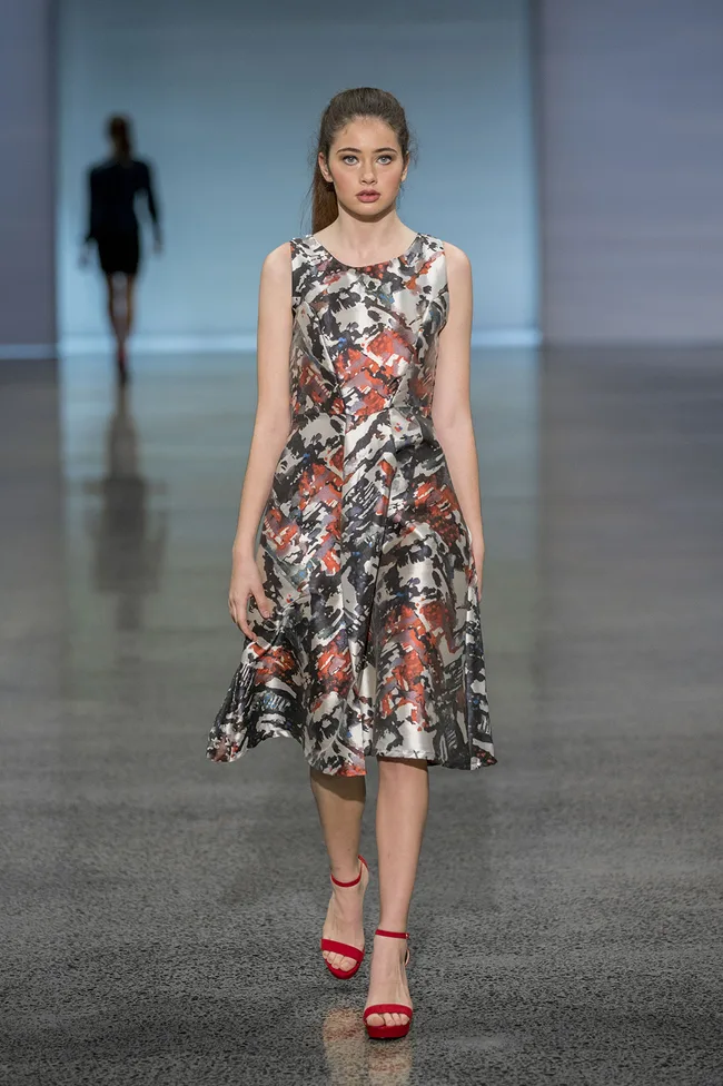 Model in a patterned dress and red heels walks down the runway, with another model blurred in the background.
