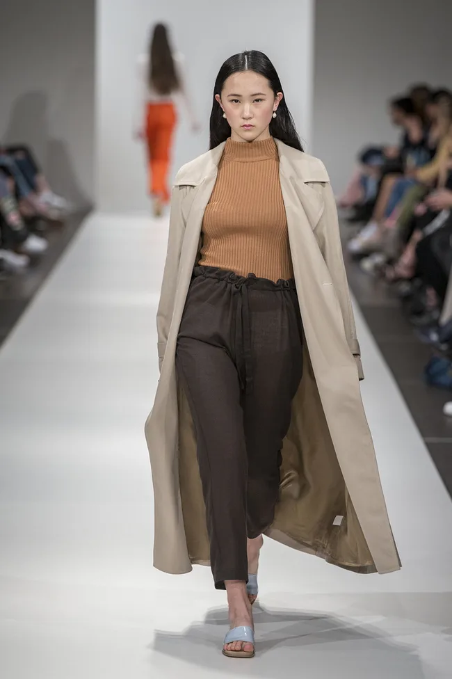 Model on runway wearing beige coat, brown turtleneck, and dark pants; audience seated along the runway.