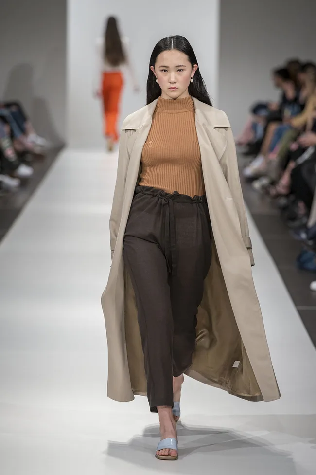Model on runway wearing beige coat, brown turtleneck, and dark pants; audience seated along the runway.