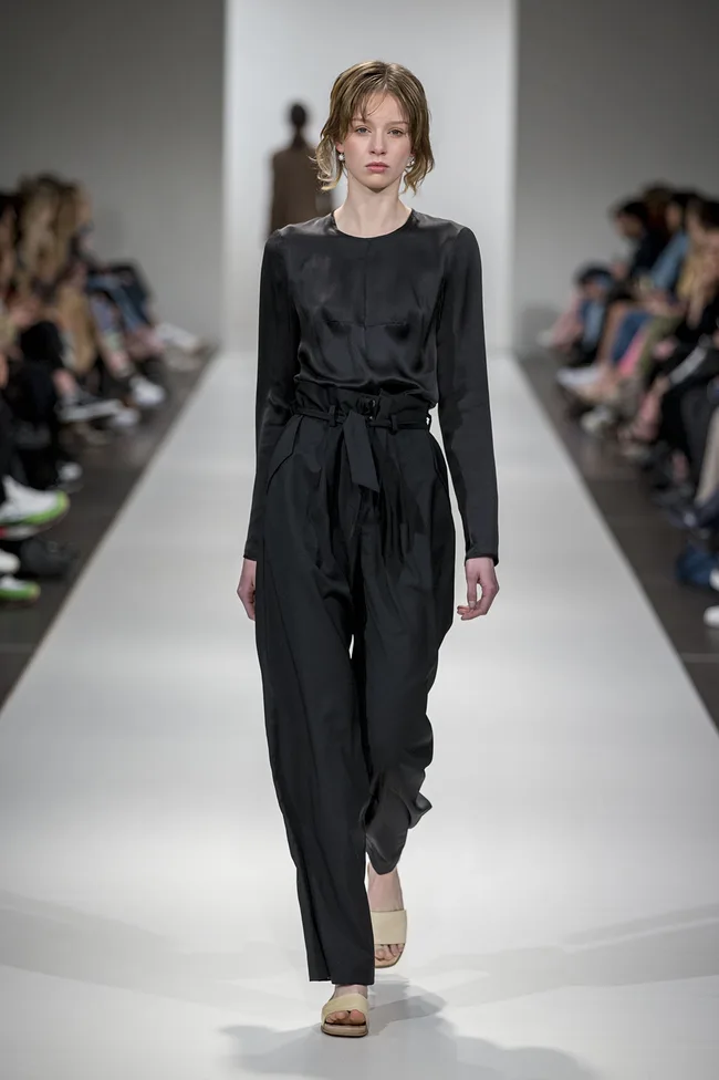 Model in black outfit walking on runway during a fashion show, surrounded by audience.