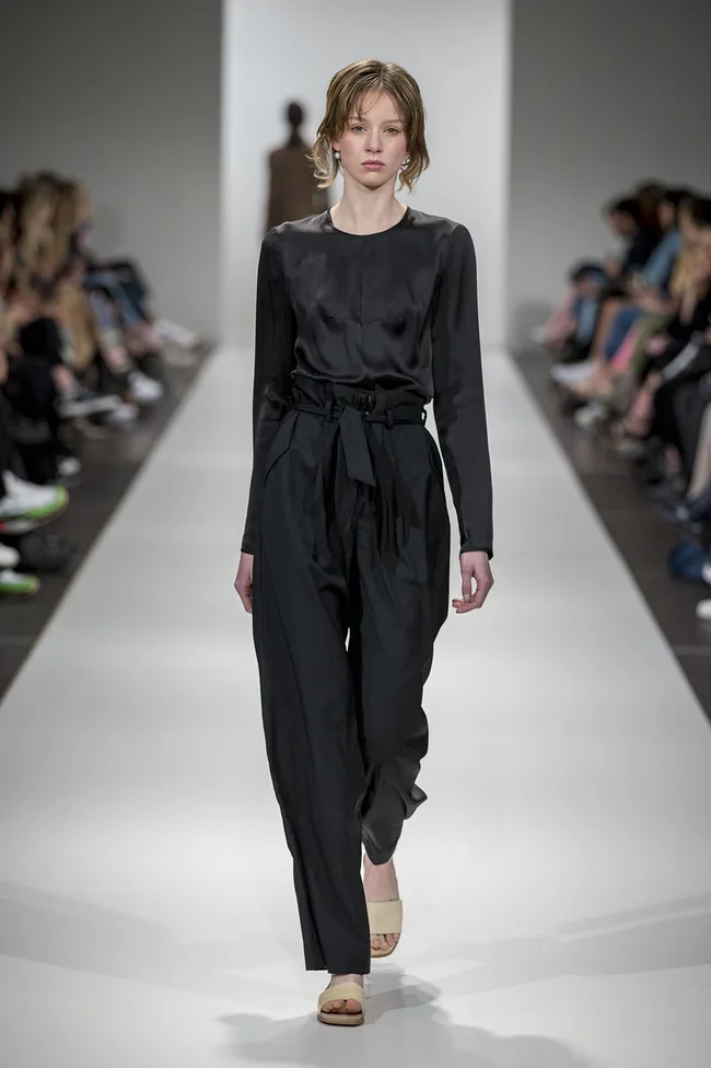 Model in black outfit walking on runway during a fashion show, surrounded by audience.