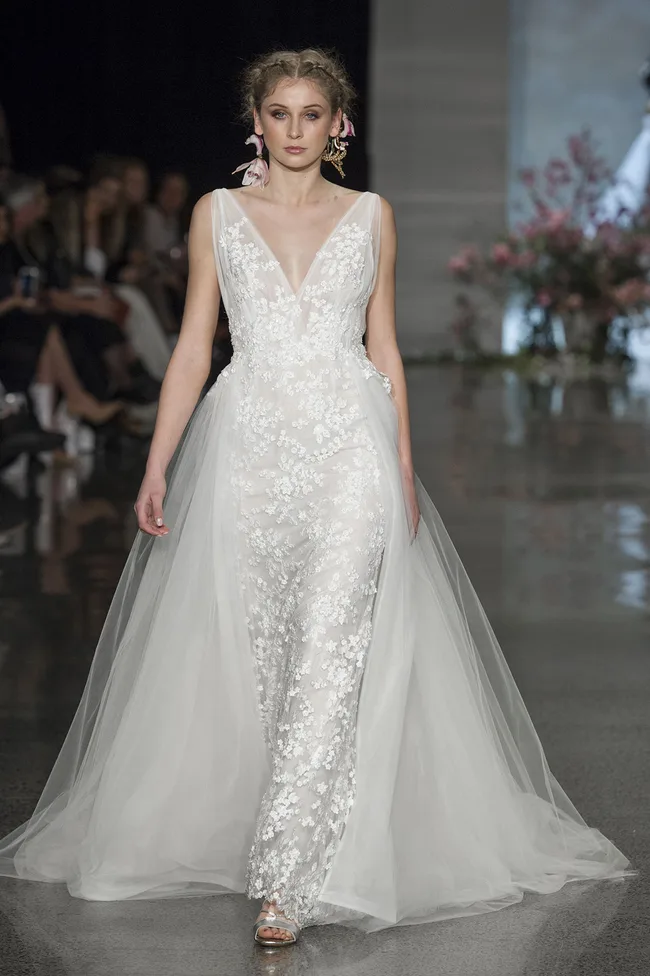 Model in a white lace wedding gown on the runway, floral details, wearing large earrings, with floral background.