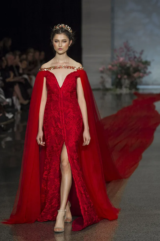 Model in an elegant red gown and crown walking down the runway at a fashion show, floral background.