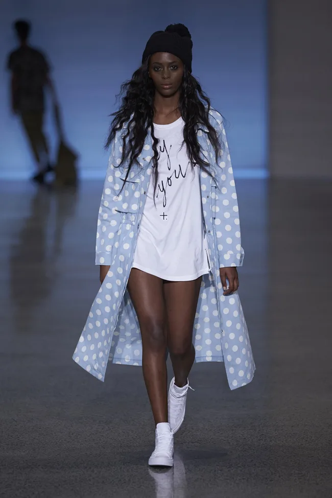 Model walking runway in polka dot coat, white shirt with text, beanie, and sneakers.