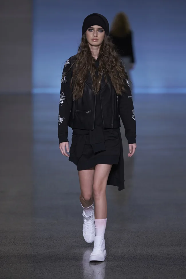 Model in black outfit with floral designs, beanie, and white sneakers walking on runway.