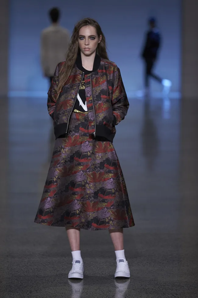 Model wearing a colorful floral bomber jacket and skirt set with white sneakers on a runway.