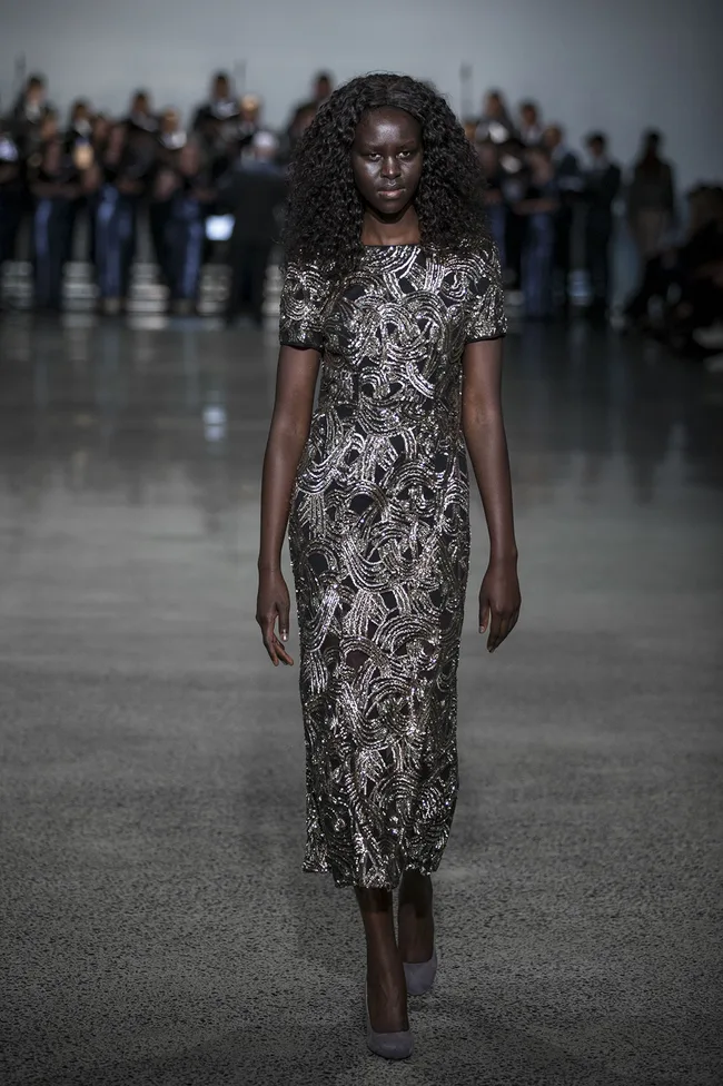 Model in a shimmering patterned dress walks the runway confidently, with a blurred audience in the background.