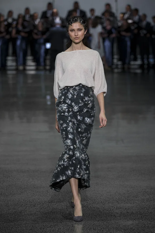 Model in a light top and floral skirt walks runway, with audience in the background.