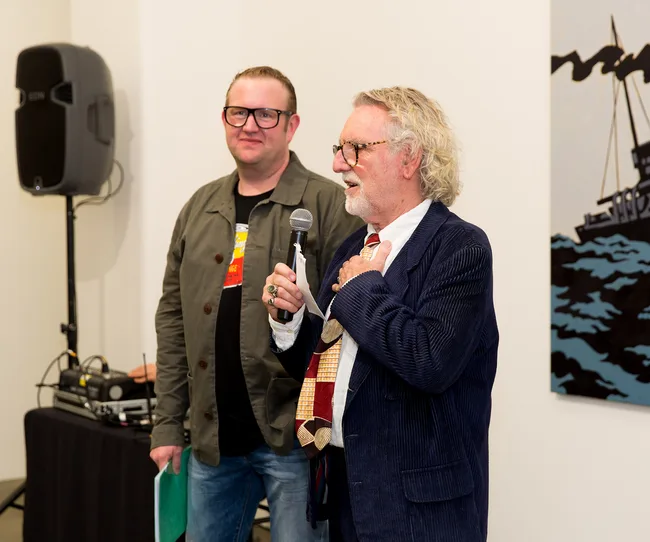 Two men standing indoors; one holding a microphone and speaking, with a speaker and artwork in the background.