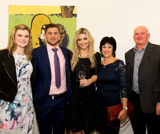 Group of five people posing in formal attire, with a colorful painting in the background. One holds a wine glass.