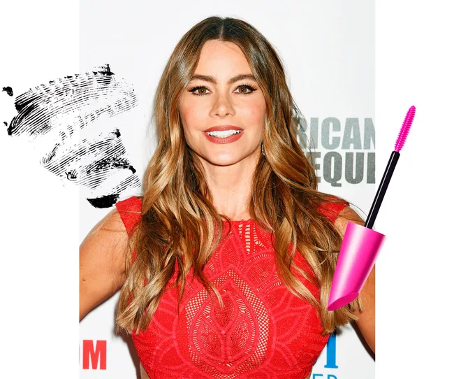 Celebrity with wavy hair in a red dress, smiling, with mascara and brush next to her head on a white background.
