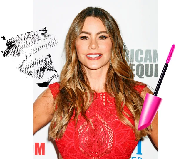 Celebrity with wavy hair in a red dress, smiling, with mascara and brush next to her head on a white background.