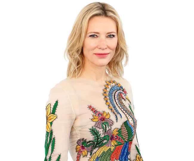 Blonde woman in a floral and peacock embroidered dress, smiling slightly against a white background.