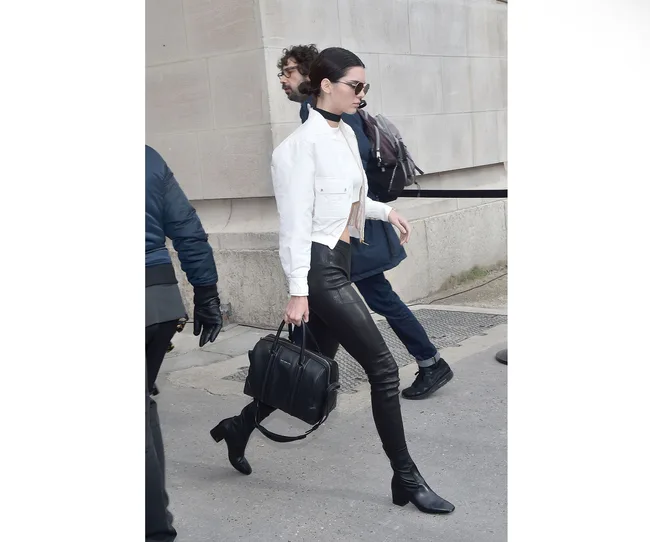 Walking woman in white jacket, black leather pants, black boots, sunglasses, carrying a handbag.