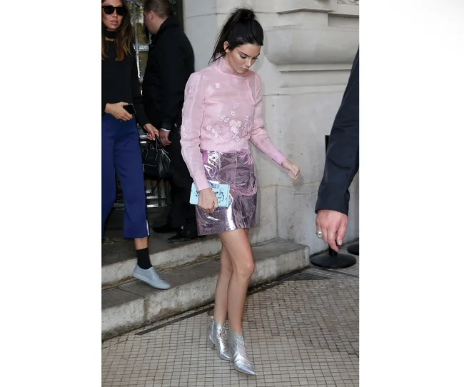 A person in a pink top and shiny skirt with silver boots, holding a small bag, walking on a tiled surface outside a building.