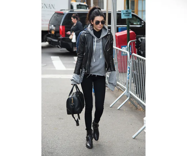 Woman in a leather jacket and sunglasses walking on a city street, carrying a black handbag.