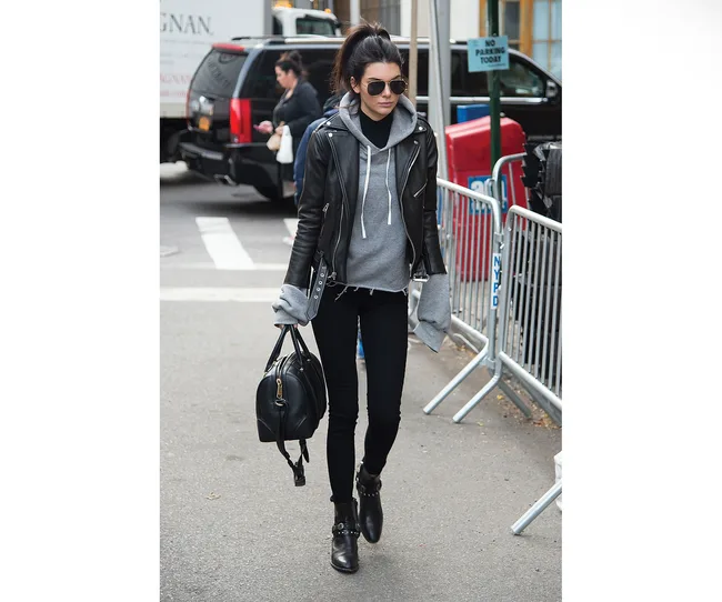 Woman in a leather jacket and sunglasses walking on a city street, carrying a black handbag.