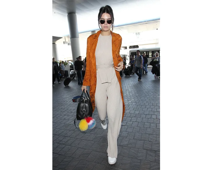 Woman in long orange coat, beige outfit, sunglasses, carrying a black bag with colorful pom-pom, walking in airport.