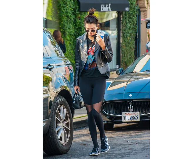 A woman in a leather jacket and sunglasses walks with a drink beside parked cars on a street.