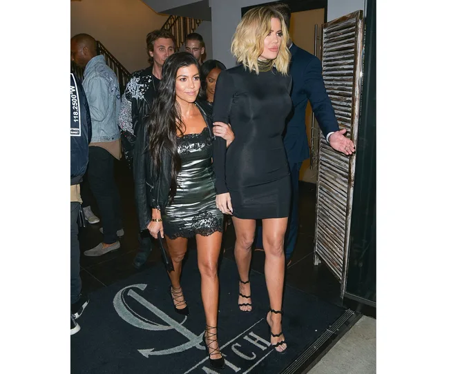 Women in stylish black outfits walk together in a crowded room, appearing to leave an event.