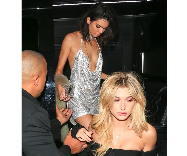 Two women stepping out of a car at night, one in a sparkly dress, the other in a black outfit, guided by an assistant.