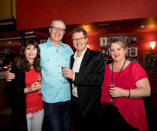 Four people standing together in a bar, holding glasses of champagne, smiling at the camera.