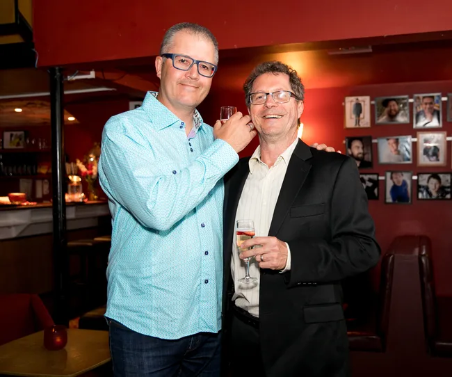 Two men smiling and holding drinks in a warmly lit room with framed photos on the wall.