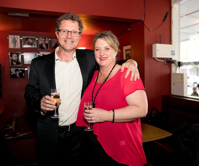 A man and woman smiling, holding champagne glasses in a warmly lit room with framed photos on the wall behind them.