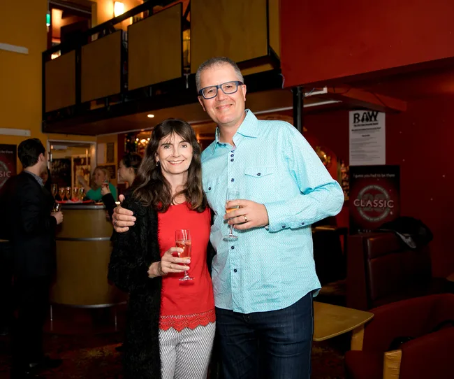 A man and woman smiling, holding drinks in a cozy bar setting.
