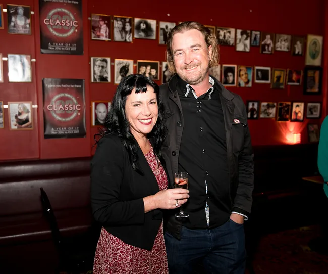 Two people smiling indoors, one holding a glass, with framed photos on a red wall in the background.