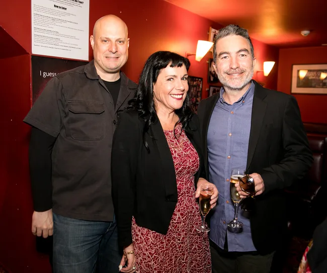 Three people smiling in a red room; two men and one woman holding glasses of drinks, dressed in semi-formal attire.