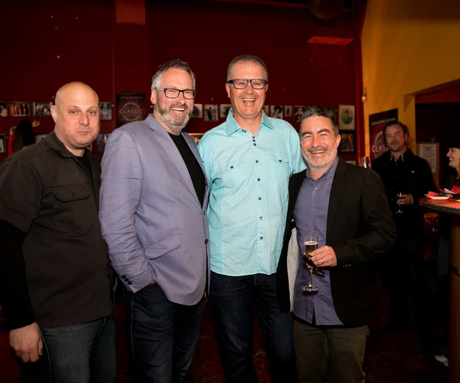 Group of four men smiling and posing together at an indoor event, with drinks in hand.