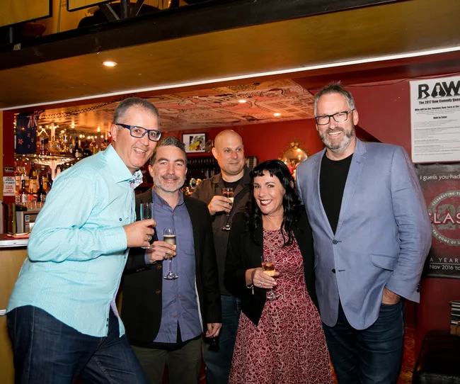 Group of five people smiling and holding drinks in a warmly lit indoor setting.