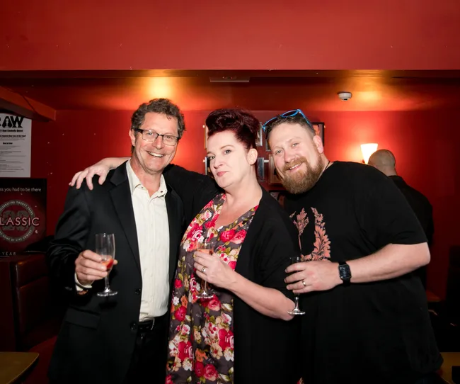 Three people smiling and holding drinks, posing in a warmly lit room.