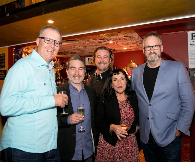 A group of five people smiling, holding drinks at a social gathering in a warmly lit room.