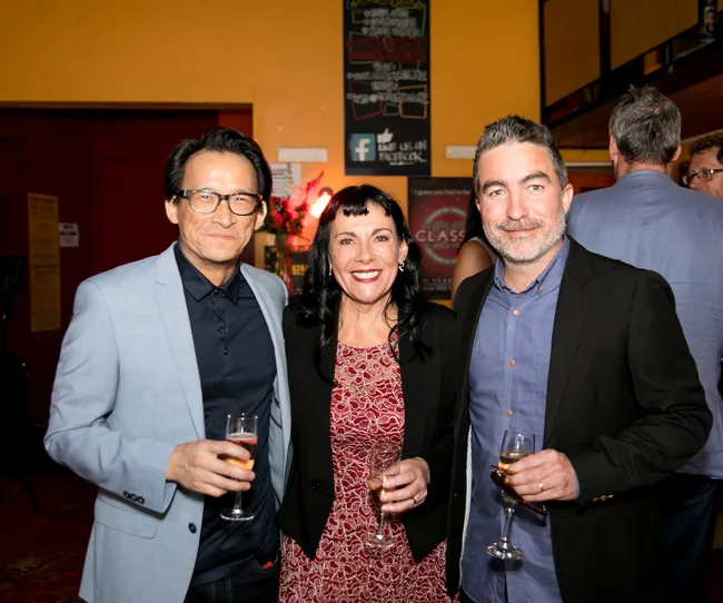 Three people smiling, holding champagne glasses at an indoor event with a yellow background and wall posters.