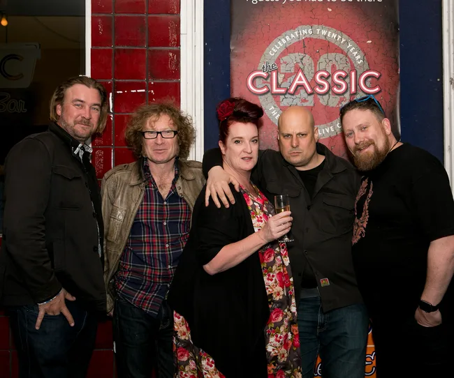 Group of five people posing in front of "The Classic" sign on a red-tiled wall, one holding a drink.