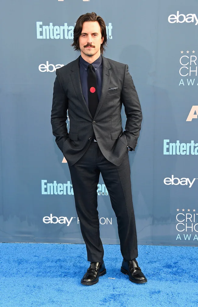 A man in a dark suit and tie stands on a blue carpet at an awards event, with logos in the background.