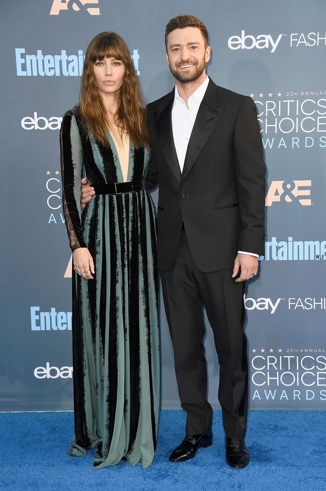Couple posing on blue carpet at the Critics' Choice Awards, woman in velvet gown, man in black tuxedo, smiling.
