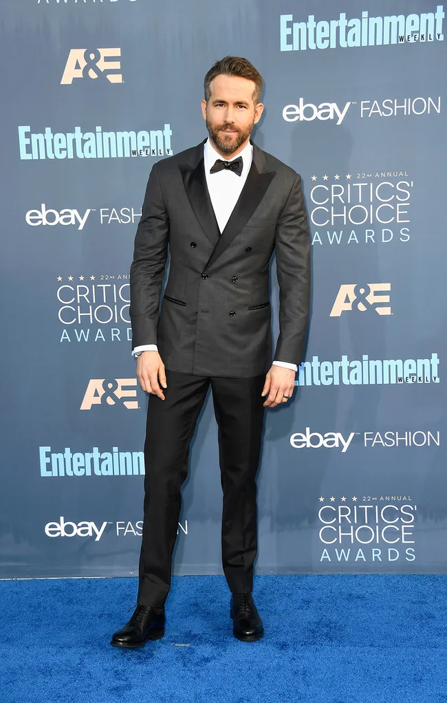 Man in a tuxedo on a blue carpet at the Critics' Choice Awards with a backdrop of sponsor logos.