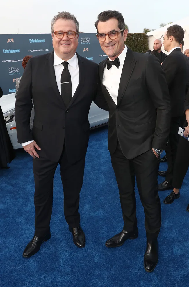 Two men in suits pose on a blue carpet at an awards event with "Critics Choice Awards" signage in the background.