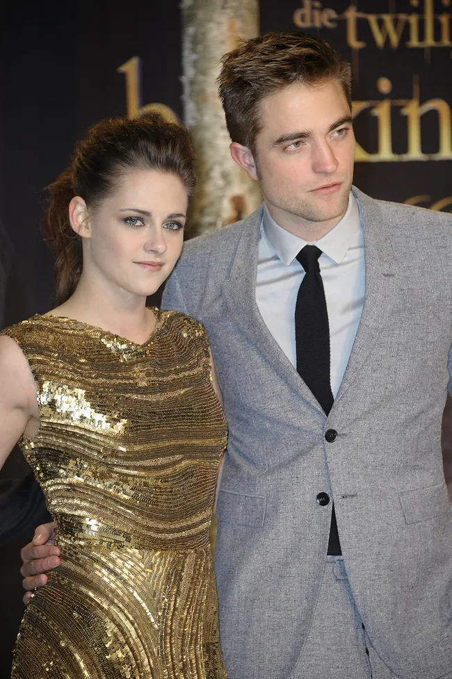 Pair posing together on red carpet; woman in gold dress, man in gray suit with black tie; Twilight-themed backdrop.