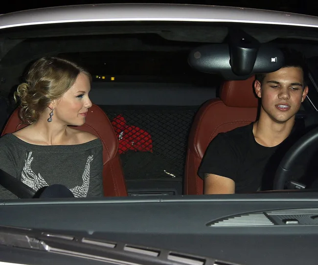 Two people sitting in the front seats of a car at night, one is driving, the other is looking at the driver, both are smiling.