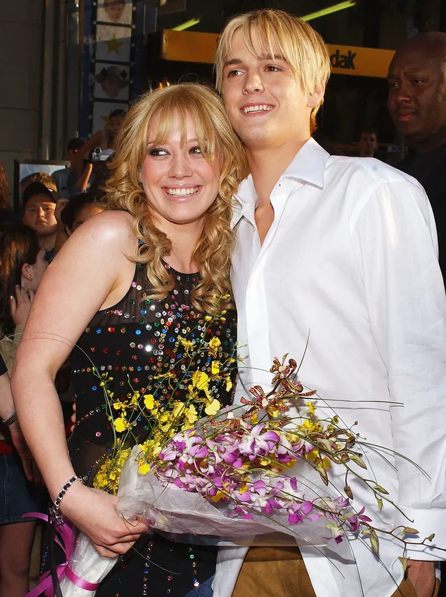 A smiling couple poses together, with the woman holding a bouquet of flowers, in an outdoor event setting.