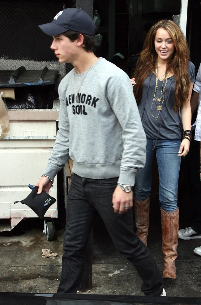Two people dressed casually walking outside, one in a "New York Soul" sweatshirt and cap, the other in boots and layered necklaces.